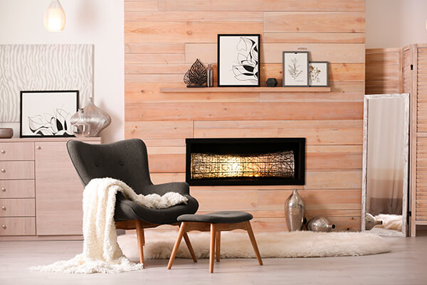 Wooden backdrop to modern fireplace with white fluffy rug in front and grey armchair and footstool